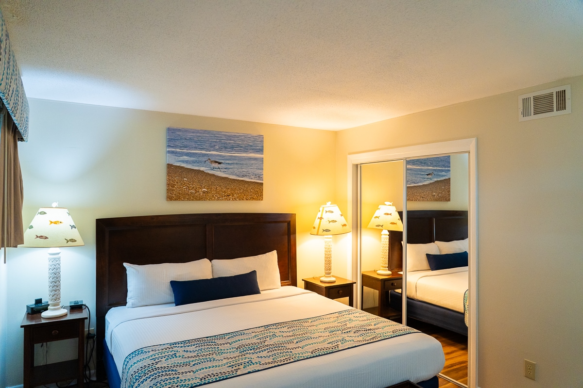 The bedroom features a spacious king-size bed with a blue decorative accent pillow, framed by two bedside lamps. A serene beach-themed artwork hangs above the bed, while mirrored closet doors reflect the soft lighting, creating a cozy atmosphere.