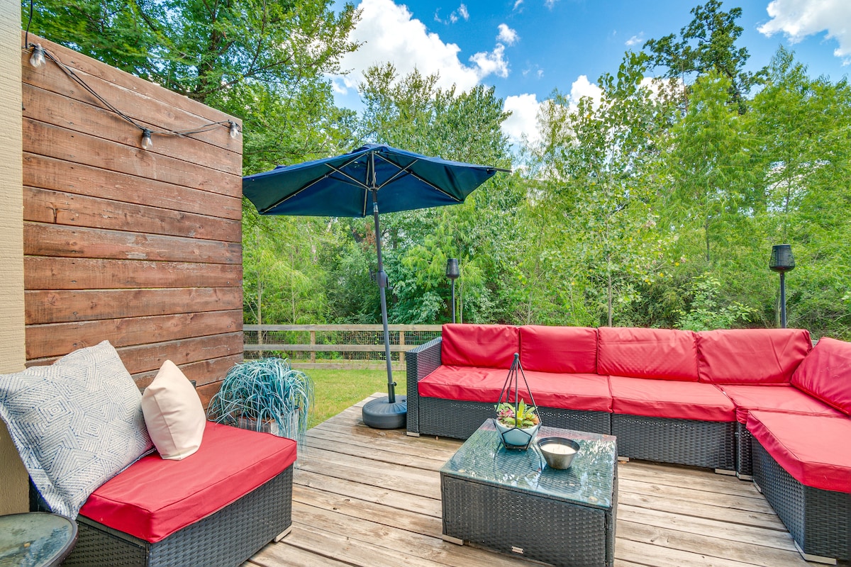 A private patio is equipped with red cushioned seating arranged around a glass-top table. An umbrella provides shade, while trees and greenery surround the space, offering a natural view. String lights are visible above, enhancing the outdoor atmosphere.