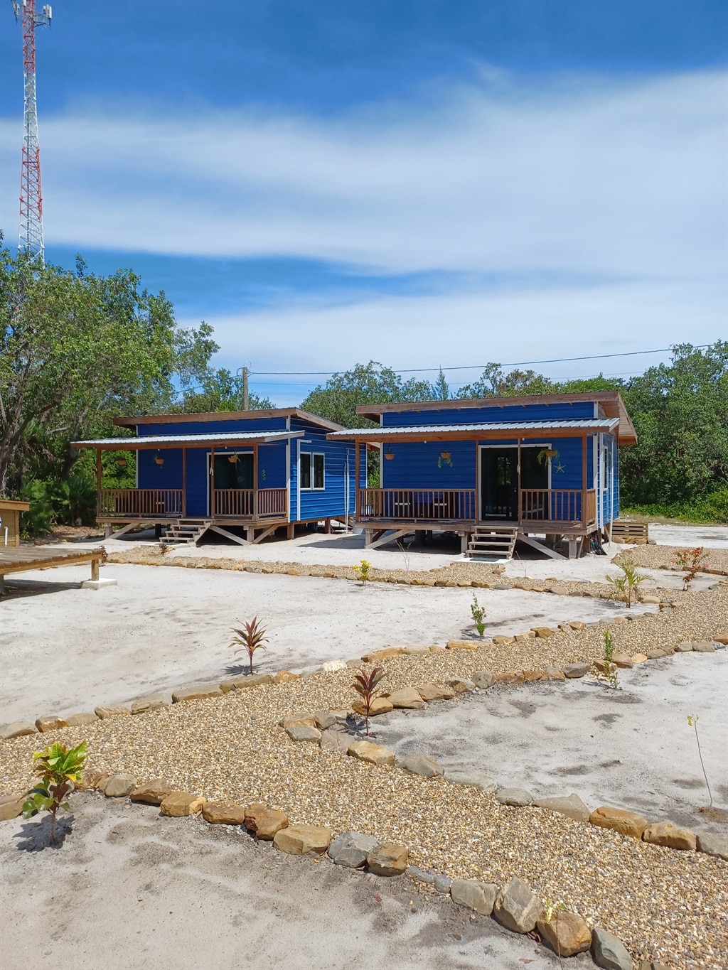 Two blue cabanas are situated on a sandy area, elevated on wooden platforms. Each cabana features a porch with steps leading down to the surrounding landscape. A landscaped garden, with a winding stone path and small plants, is visible in the foreground, enhancing the natural setting.