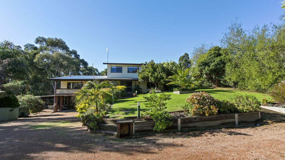 A large house is set amidst landscaped gardens and lush greenery. A gravel pathway leads to the front entrance, flanked by various shrubs and trees. The two-story home features a spacious design with a balcony and outdoor area visible on the upper level.