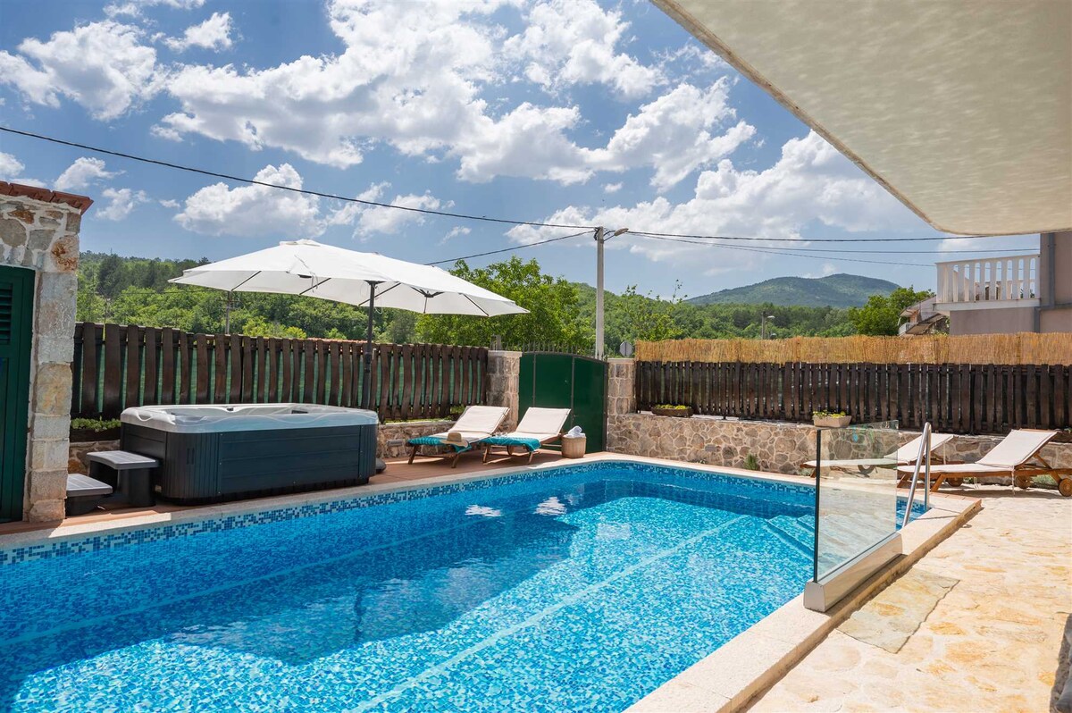 An inviting outdoor area features a swimming pool surrounded by a stone deck. A Jacuzzi is positioned beside the pool. Sun loungers are placed for relaxation, with an umbrella providing shade. Lush green hills and a clear sky complete the serene setting.