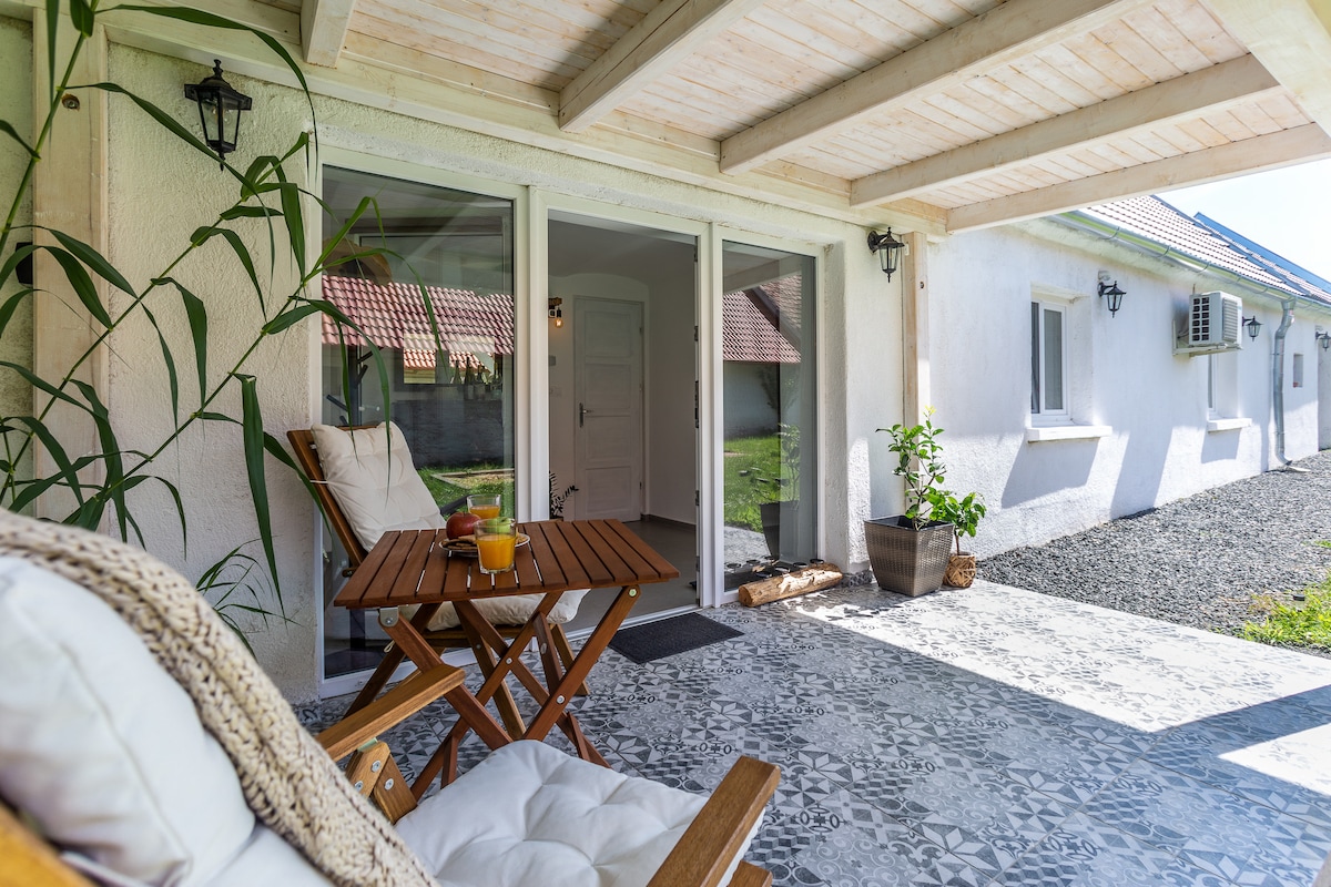 A shaded porch is adorned with wooden deck chairs and a small table, inviting relaxation. The entrance features a glass door leading into the elegant hallway, while decorative plants enhance the welcoming ambiance.