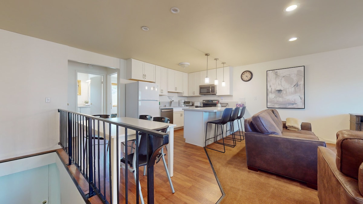 The open-plan living and dining area displays a modern kitchen with white cabinetry and stainless steel appliances. A dining table for six is visible, complemented by three bar stools at the kitchen counter. A cozy seating arrangement with a brown leather couch invites relaxation, with soft lighting enhancing the space.