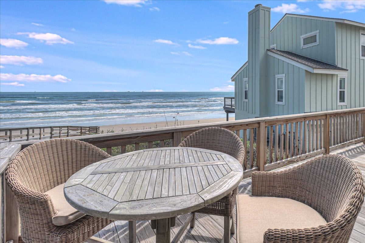 An outdoor seating area is positioned on a spacious balcony, featuring a round wooden table surrounded by two woven chairs. The ocean view stretches in the background, with gentle waves and a clear sky adding to the tranquil atmosphere.