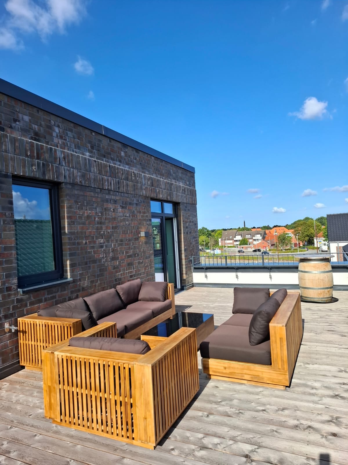 A spacious outdoor terrace is furnished with comfortable seating, featuring wooden chairs and a coffee table. The area is surrounded by a wooden railing, providing a clear view of the surrounding landscape and blue sky, enhancing the inviting atmosphere for relaxation.