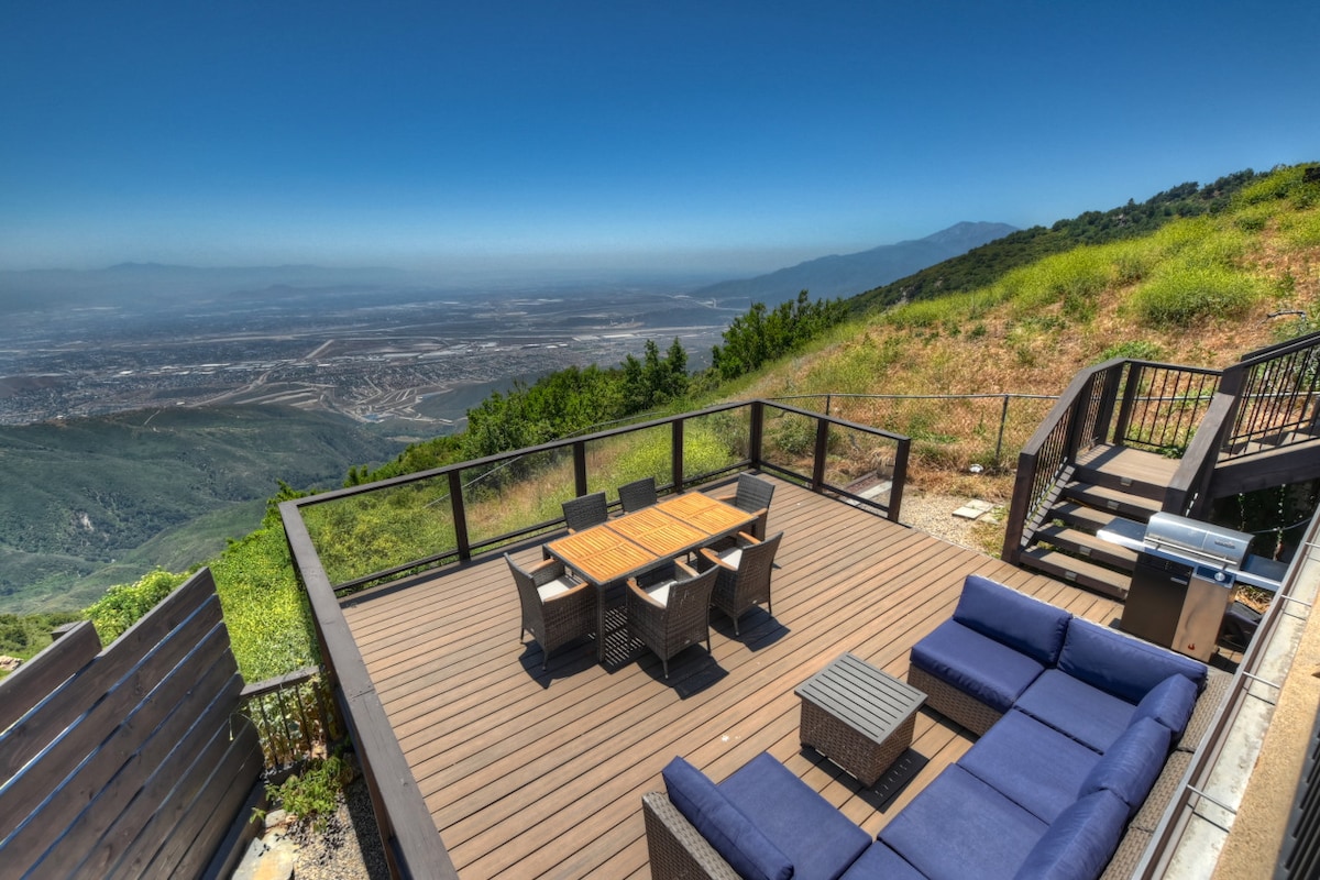 An outdoor deck features a dining table surrounded by six chairs and a sectional sofa with deep seating, all facing expansive views of the valley below. Lush greenery lines the backdrop, and clear skies enhance the visibility of the landscape.