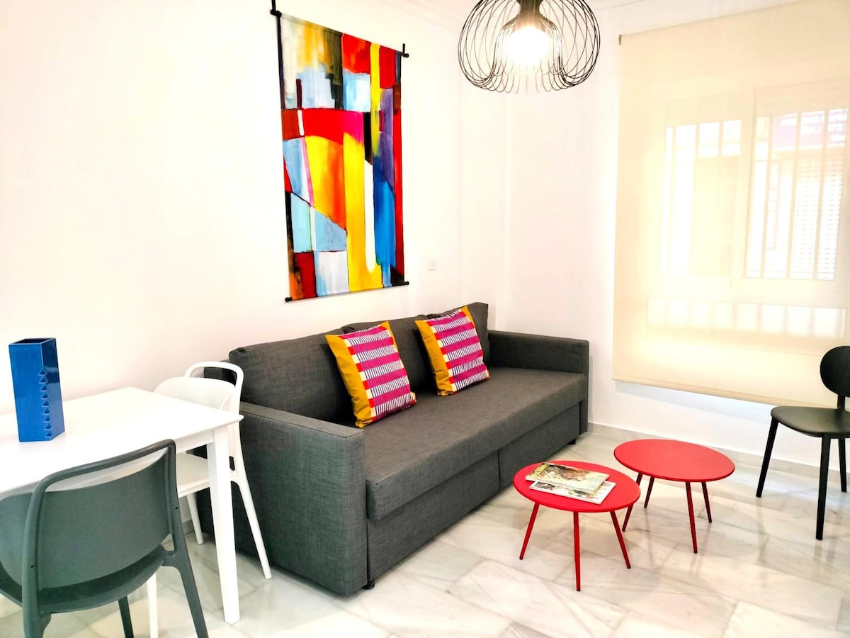 A living area features a gray sofa adorned with striped cushions in vibrant colors. Two red coffee tables are positioned in front of the sofa, while a dining table with four chairs is nearby. A colorful wall art piece adds interest, along with a softly lit window.
