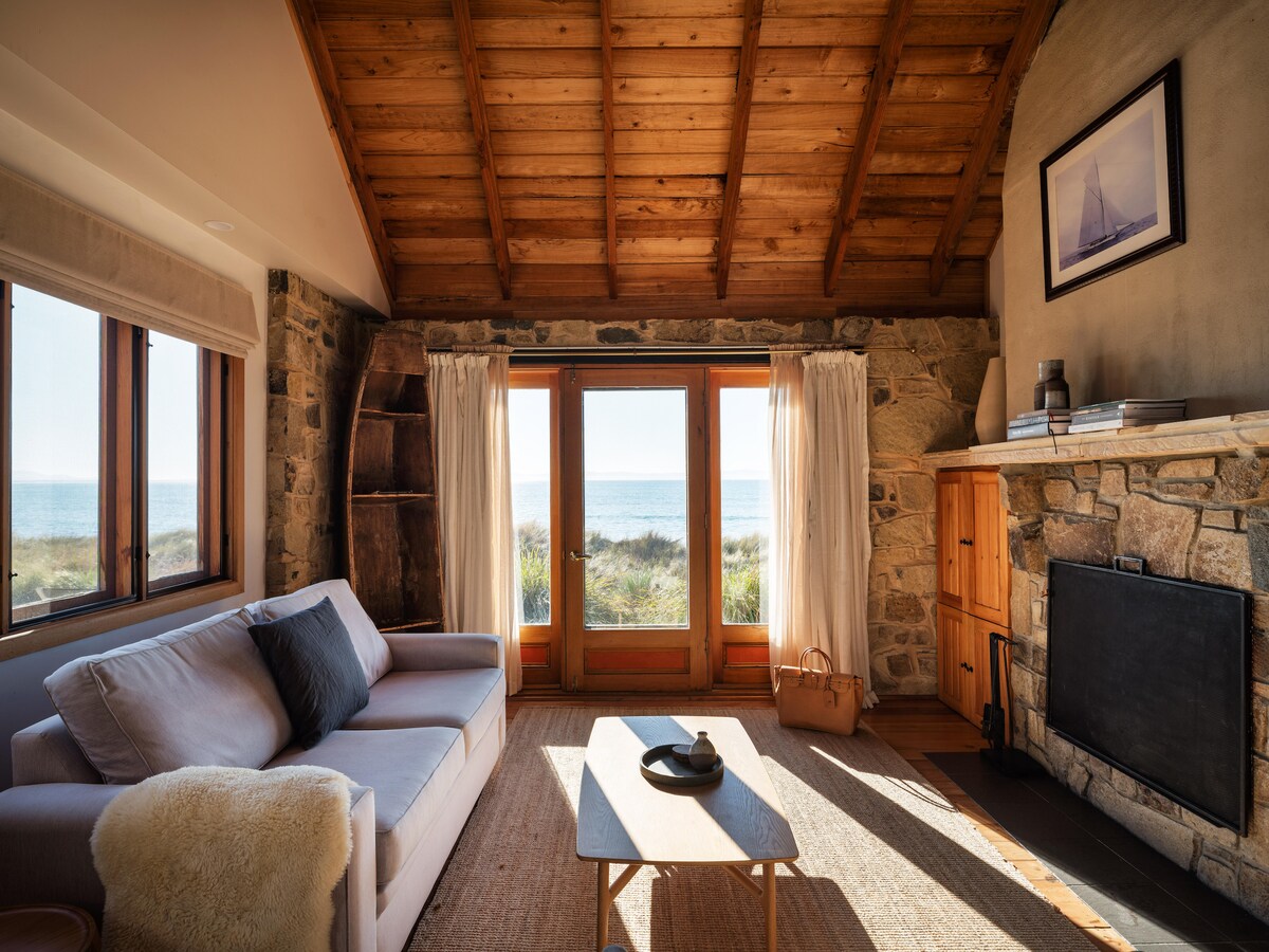 A bright living area features large windows with views of the water, allowing natural light to fill the space. A comfortable sofa is positioned next to a minimalistic coffee table on a soft area rug. A stone fireplace stands opposite the seating area, enhancing the rustic charm.