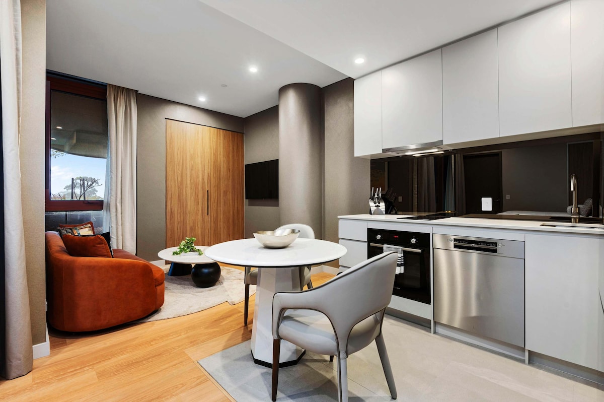 A modern kitchen area features sleek cabinetry and stainless steel appliances, including an oven and dishwasher. A round dining table is set with a simple centerpiece. Nearby, a cozy seating space is highlighted by a rust-colored armchair, contributing to a welcoming ambiance.