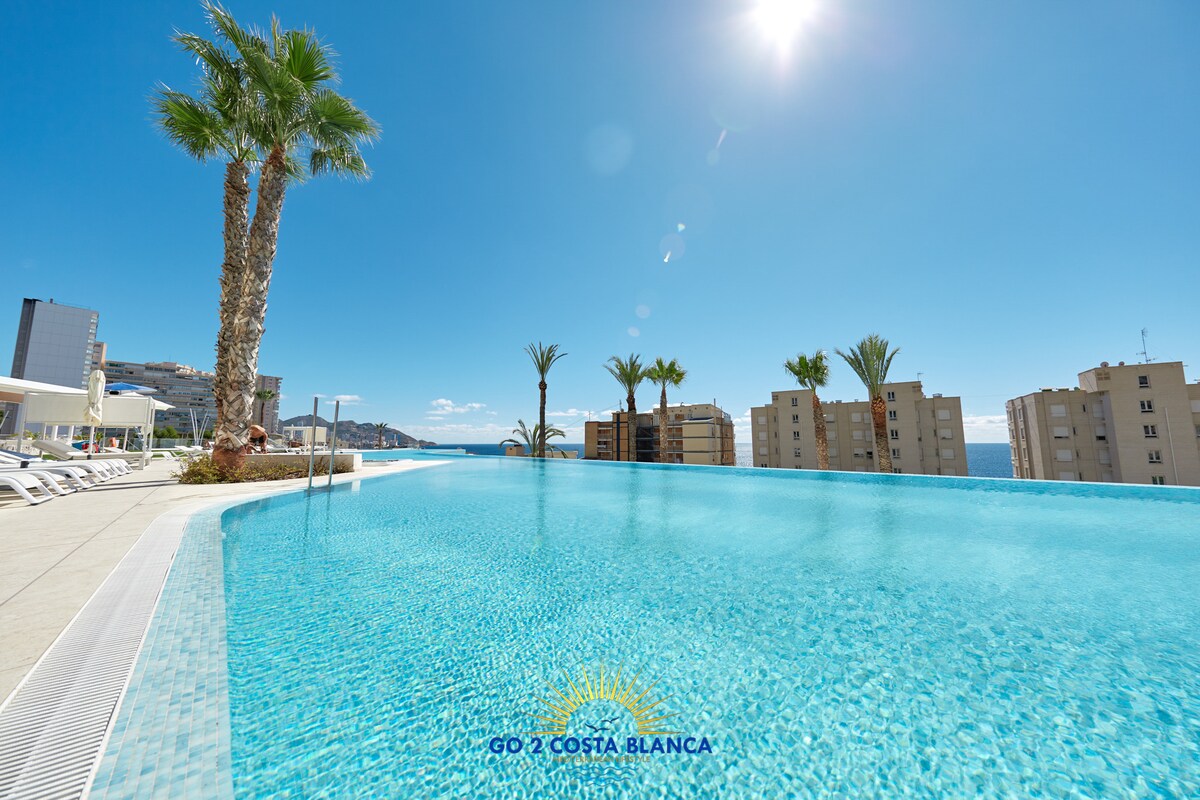 An expansive infinity pool is featured, overlooking the landscape. Palm trees line the edges, and the clear blue sky reflects in the shimmering water. Surrounding buildings are visible in the distance, enhancing the sense of relaxation by the pool area.