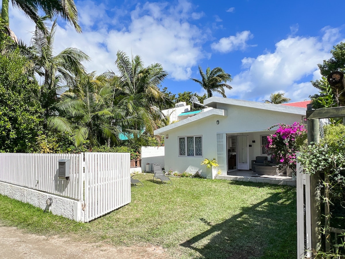 The exterior of the house is surrounded by lush greenery, including palm trees and vibrant flowers. A white wooden fence encloses the front yard, which features well-maintained grass. The terrace area is visible, overlooking the garden and inviting outdoor seating.