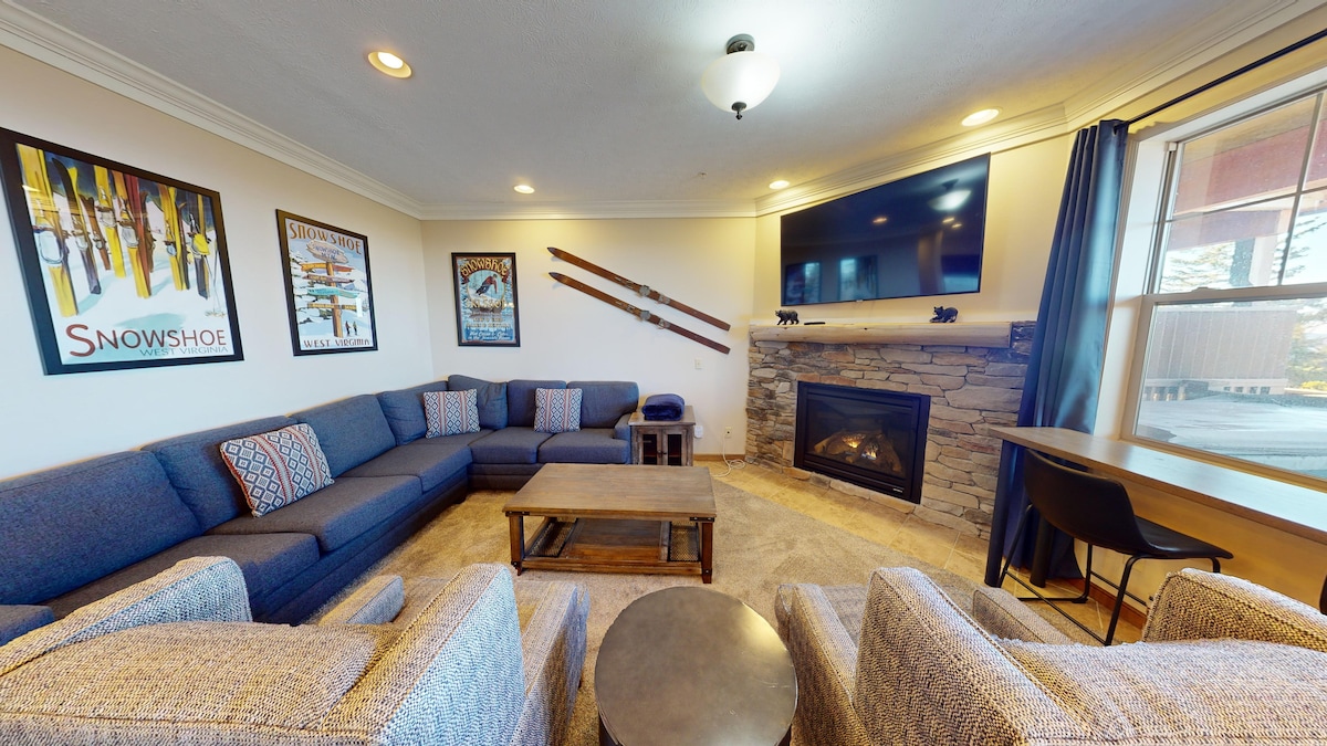 A spacious living area features a large sectional sofa, arranged around a stone fireplace. Ski-themed artwork adorns the walls, complementing the warm tones of the decor. A flat-screen television is mounted above the fireplace, and a small work desk sits near the window.