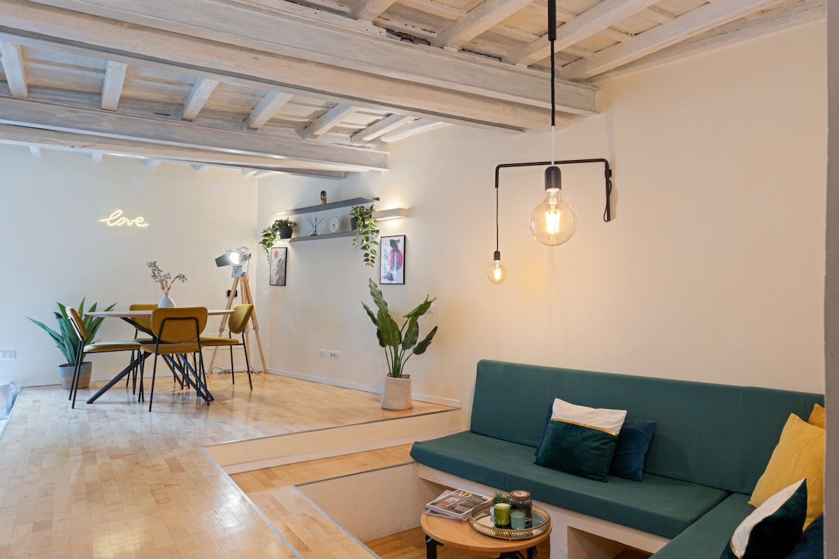 The living area features a green seating arrangement with decorative pillows, complemented by a wooden coffee table. A dining area is visible, furnished with four chairs and illuminated by stylish pendant lighting. The walls display framed artwork, and plants add a touch of greenery.