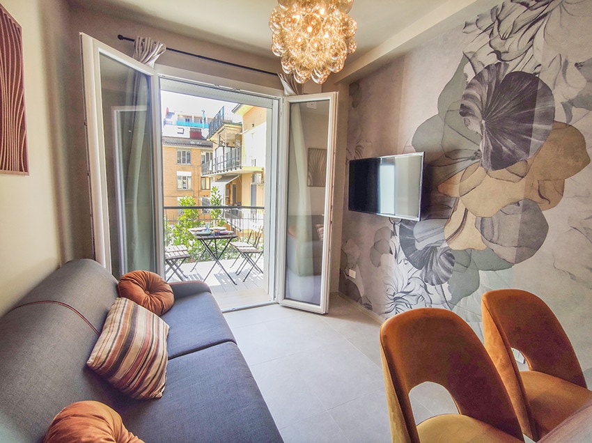 A modern living area features a comfortable gray sofa with decorative pillows and a wall adorned with soft floral patterns. Large glass doors open to a balcony with outdoor seating. A flat-screen TV is mounted on the wall, providing entertainment options.
