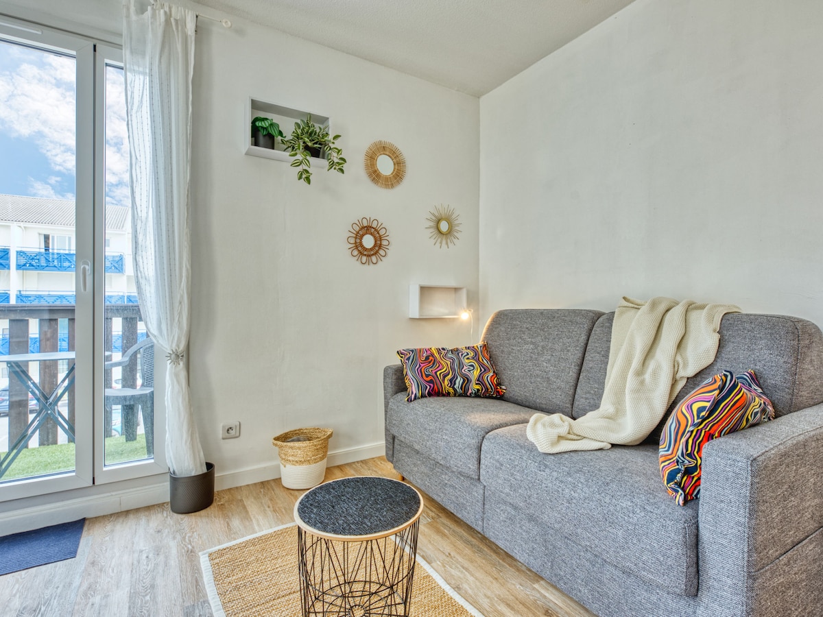 A welcoming living area features a grey sofa adorned with colorful throw pillows and a soft blanket. Natural light enters through a large window, highlighting a small round table and decorative wall elements, including mirrors and a plant, enhancing the cozy ambiance.