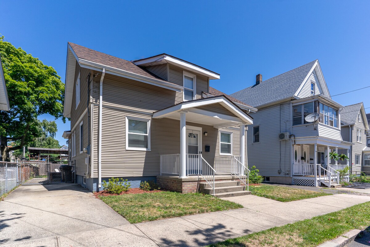 The two-story house showcases a light beige exterior, complemented by white trim and a covered front porch. A well-manicured lawn features neat flower beds along the walkway, while neighboring homes create a friendly, residential atmosphere.