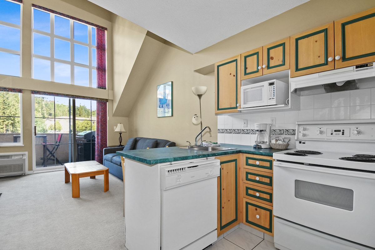 A modern kitchen area features light wood cabinetry and a contrasting dark countertop. Large windows fill the space with natural light, while a cozy seating area with a blue sofa and a coffee table is positioned nearby, creating an inviting space for relaxation.
