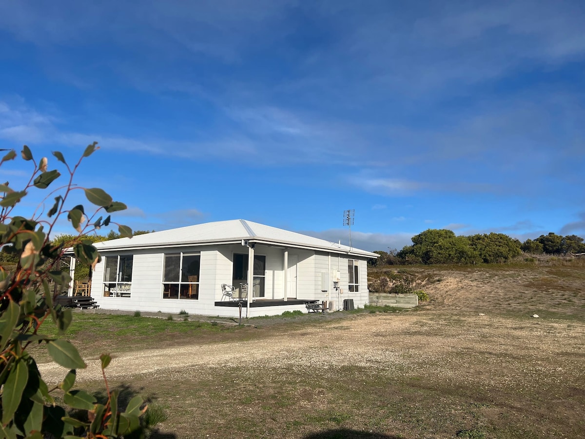 A quaint cottage is situated on three acres of land, featuring a light-colored exterior and a gabled roof. Large windows allow for ample natural light. The surrounding landscape includes gentle hills and shrubbery under a bright blue sky.