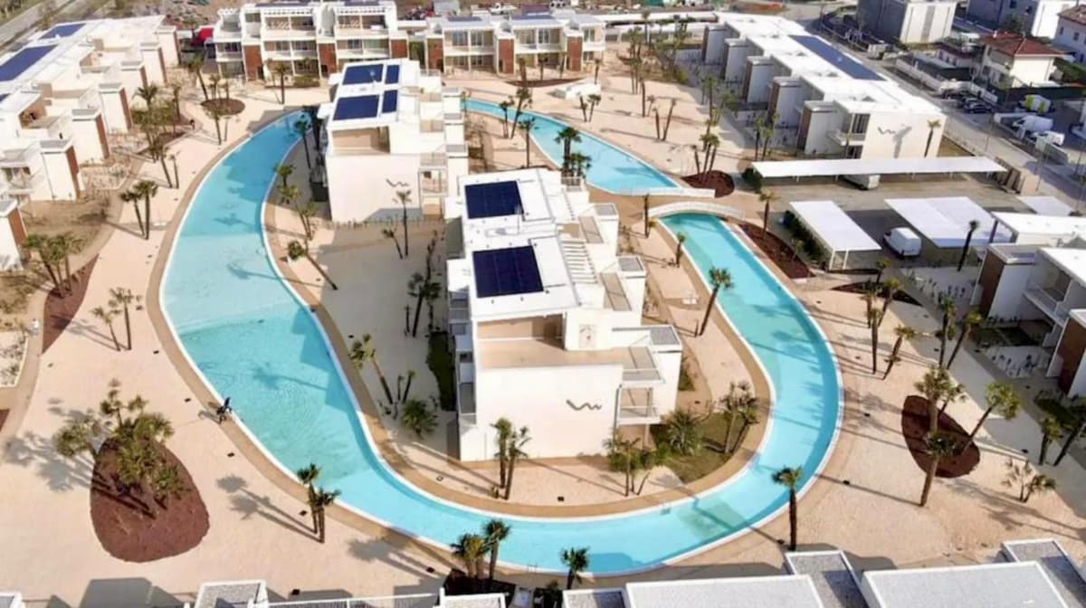 Aerial view of a modern residential complex featuring a winding pool encircled by landscaped areas. Palm trees and sandy walkways contribute to a relaxed atmosphere. Buildings arranged in a contemporary style create an open courtyard, highlighted by natural greenery and blue water.