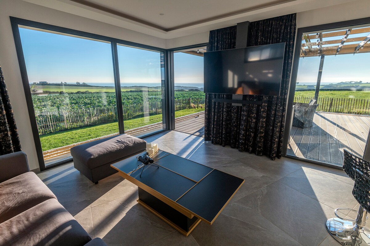 A spacious living area features large bay windows that provide expansive views of the green landscape outside. A comfortable sectional sofa and a modern coffee table are complemented by a flat-screen TV mounted on the wall. Natural light fills the room, enhancing the serene atmosphere.