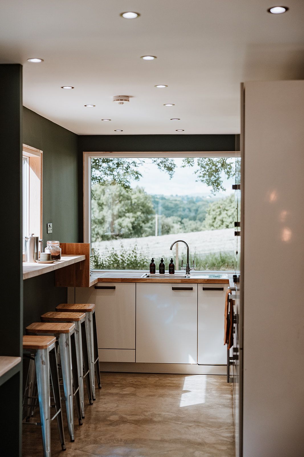 A modern kitchen features a large window offering expansive views of the surrounding greenery. Sleek cabinetry and a minimalist design are complemented by several metal stools positioned at a counter. Soft overhead lighting creates a bright atmosphere.