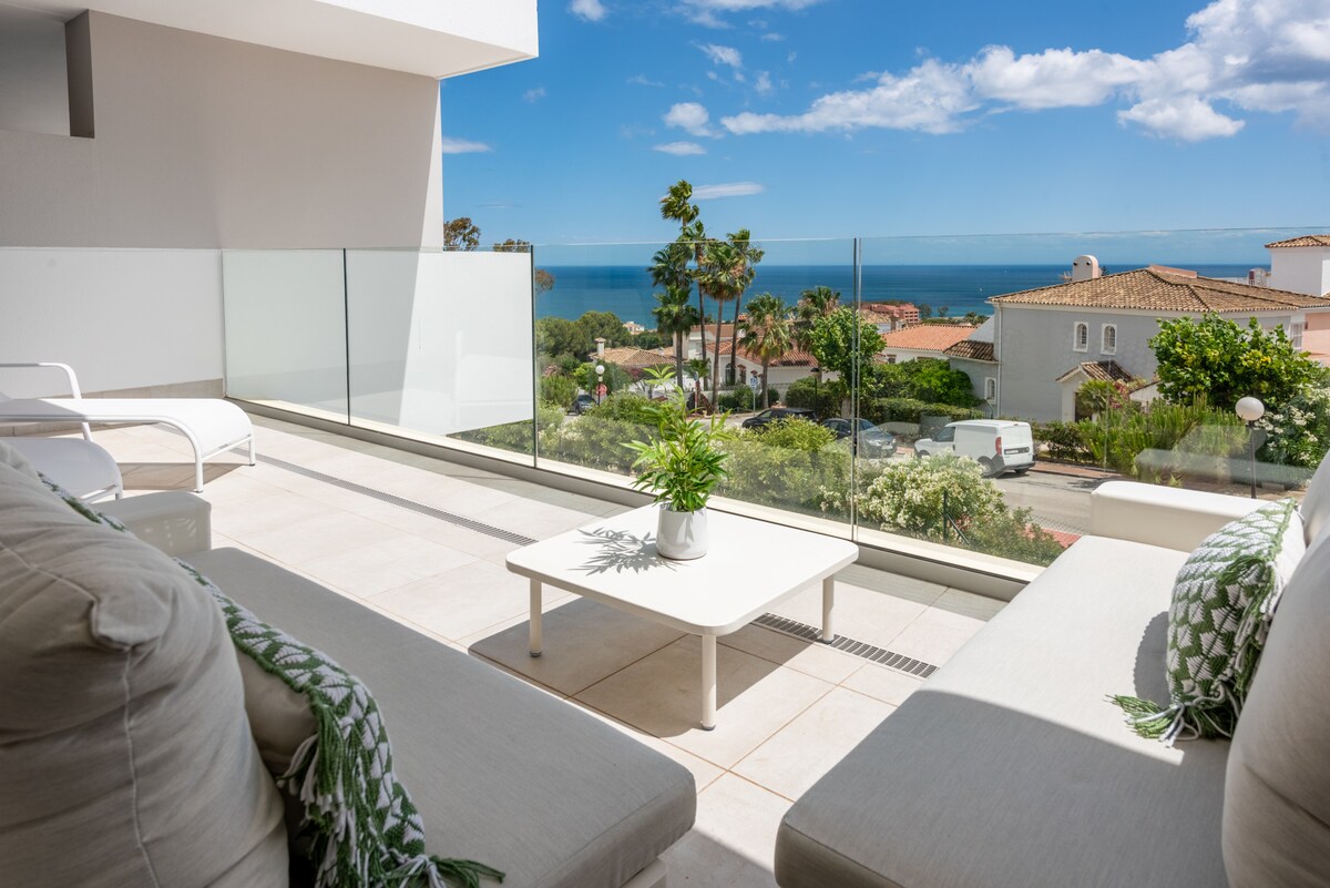 A spacious terrace features comfortable seating and a coffee table, offering unobstructed views of the sea and surrounding landscape. Glass railings enhance the openness of the space, while tropical plants add a touch of greenery, providing a relaxed outdoor environment.