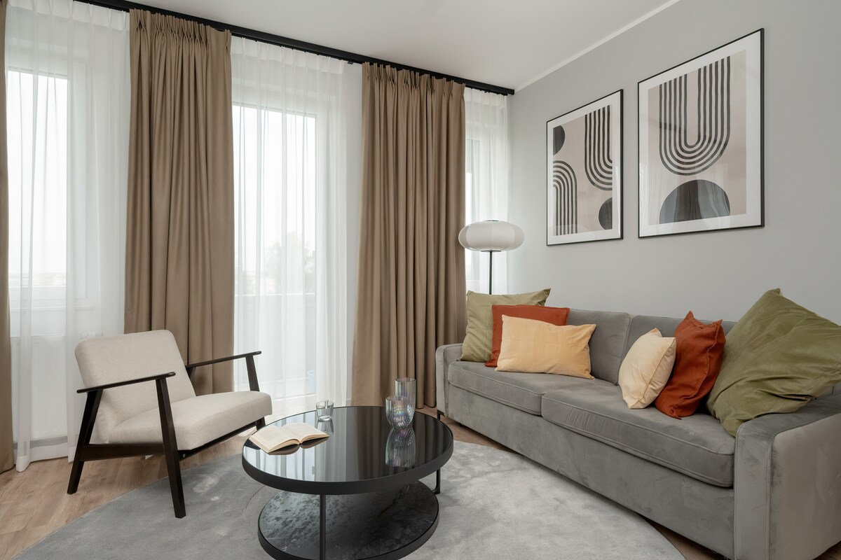 The living room features a comfortable gray sofa adorned with a mix of colorful cushions. A round black coffee table sits on a light rug, and an armchair is positioned nearby. Two framed prints decorate the wall, and large windows are dressed with sheer curtains, allowing natural light to flow in.