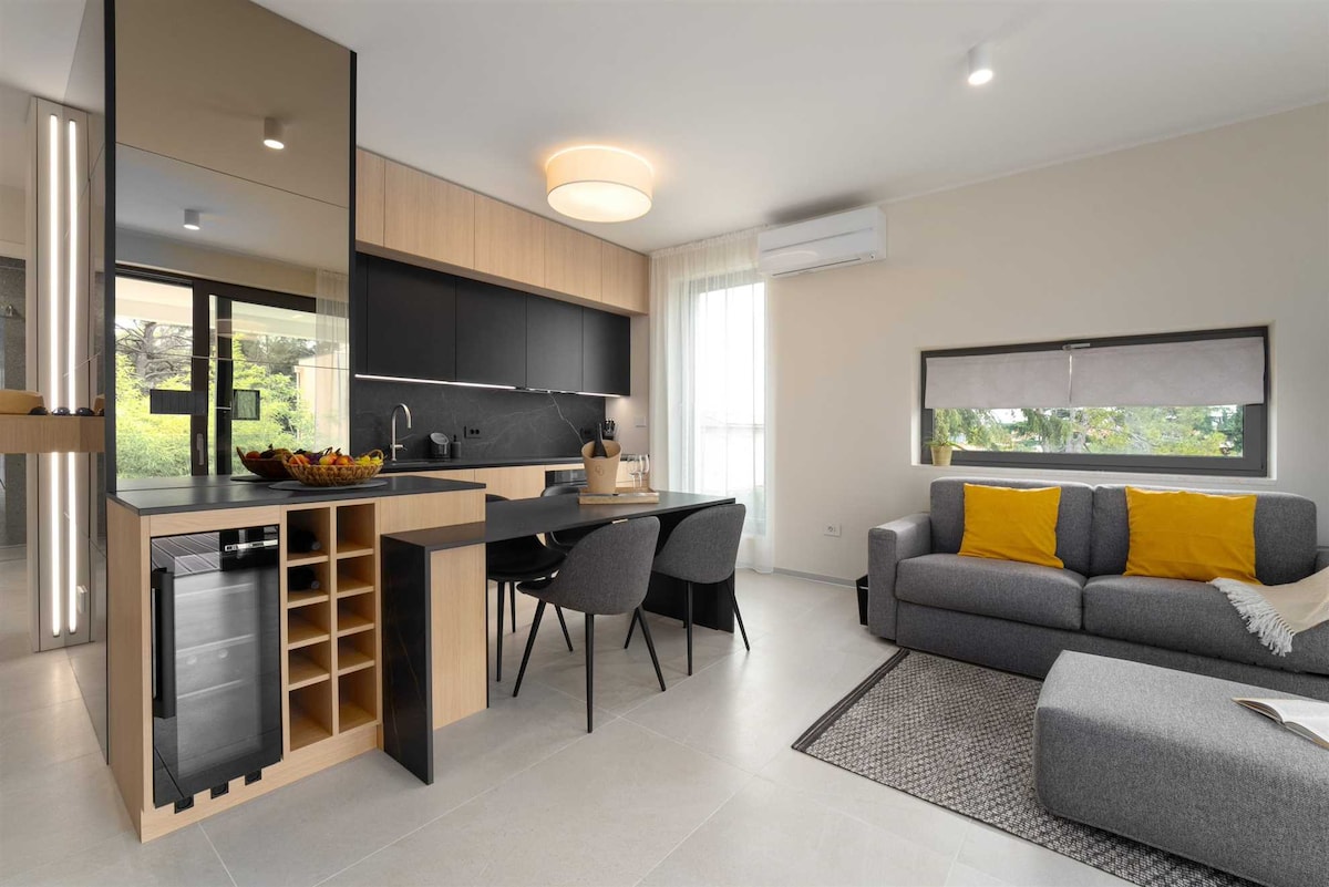 A modern kitchen and living area is presented, featuring sleek cabinetry and a dining table with four chairs. Natural light filters through large windows, illuminating a comfortable sofa adorned with yellow cushions. A small wine cooler and a textured area rug add functional and aesthetic elements.