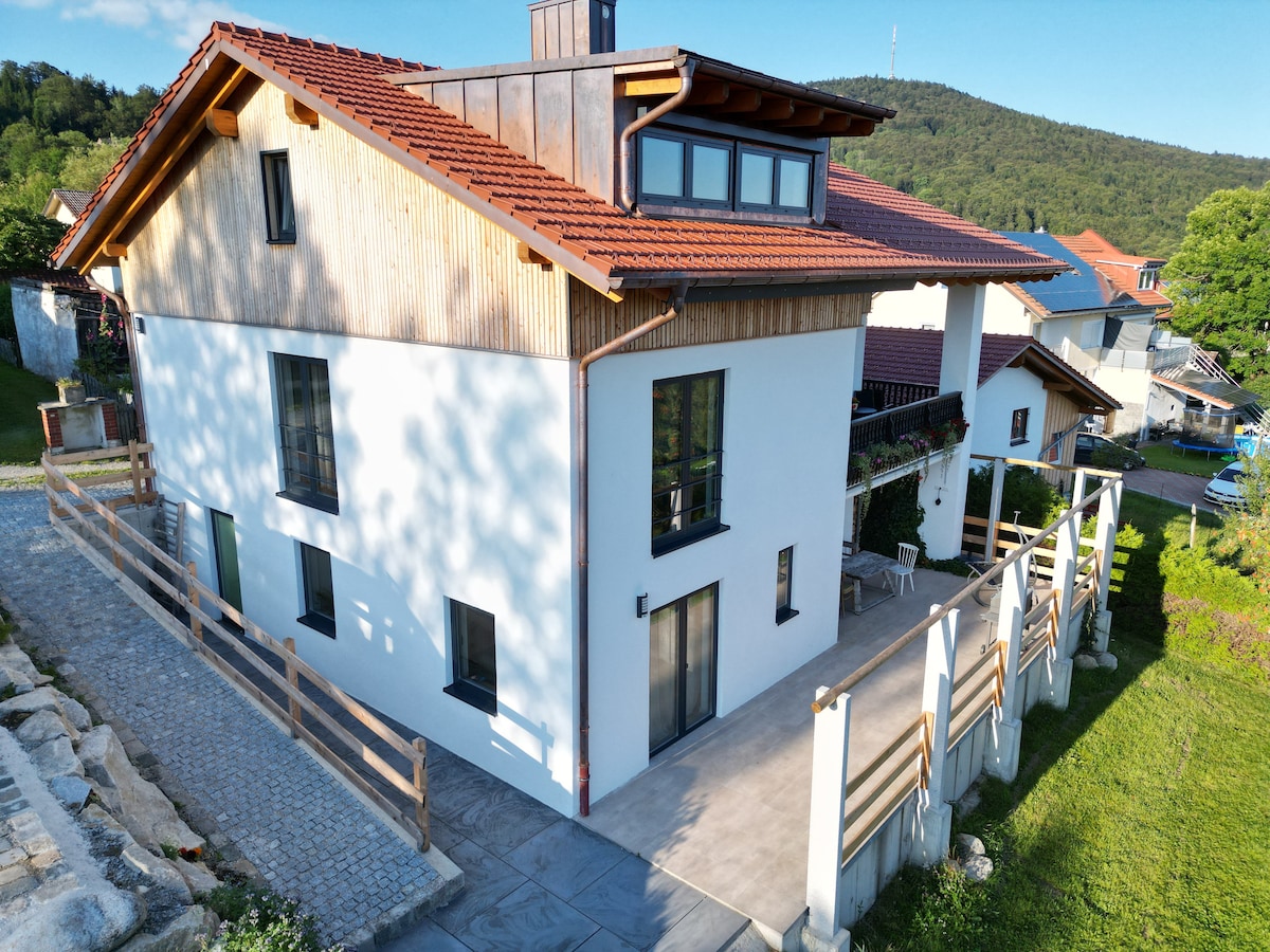 The exterior of the chalet is depicted, showcasing a contemporary design with a mix of wood and white finishes. A spacious patio area features outdoor seating, and large windows reflect natural light. Lush greenery surrounds the property, with rolling hills in the background.