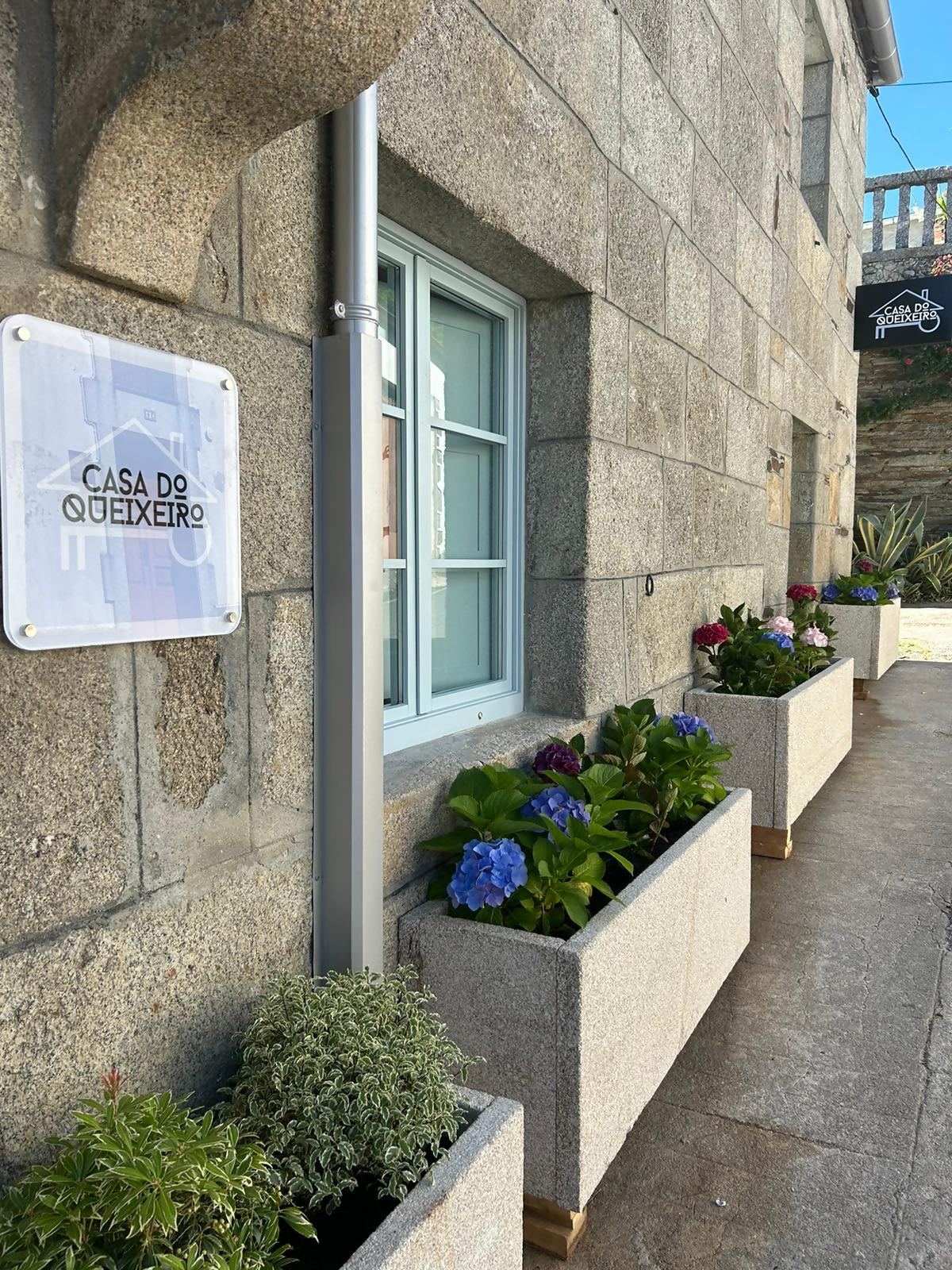 The exterior of the building features a stone facade with a clear sign identifying 'Casa do Queixeiro.' Colorful flowering plants are arranged in modern planters along the pathway, enhancing the inviting entranceway. A large window with white frames reflects natural light, contributing to the overall appeal.