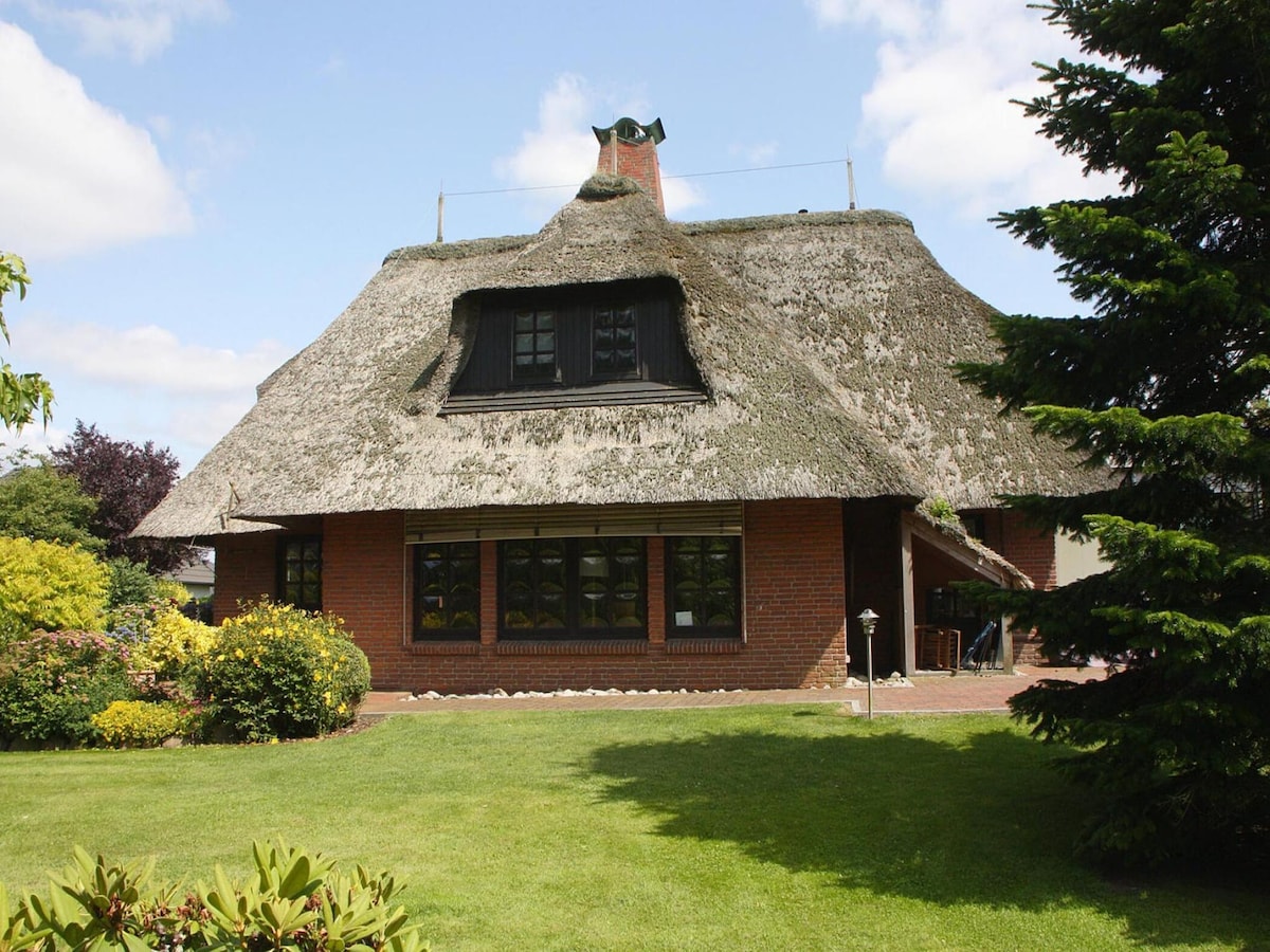 A large thatched-roof house is set amidst a green lawn, surrounded by flowering shrubs and trees. Large windows allow natural light to fill the interior, while a chimney adds a rustic touch to the charming exterior.