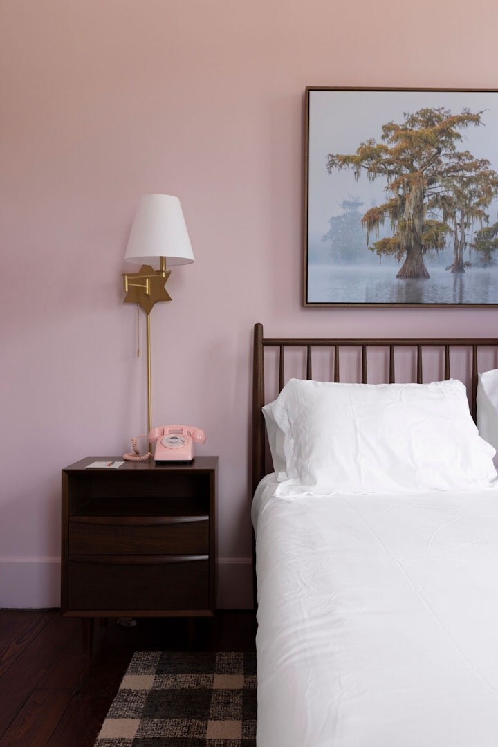 A rose-colored wall complements a wooden bed with white bedding. A sleek nightstand holds a vintage pink telephone. Natural light enhances the minimalist decor, while a framed artwork depicting serene waters and trees adds a tranquil touch.
