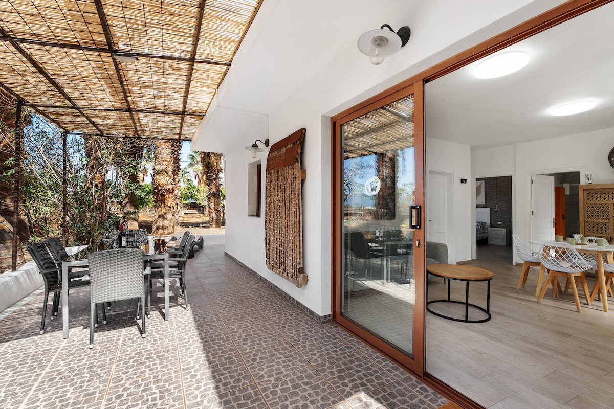 A covered terrace features a dining table with several chairs, providing an outdoor space for meals. Large glass doors offer a view into the interior living area. Natural light enhances the area, while greenery is visible in the background.