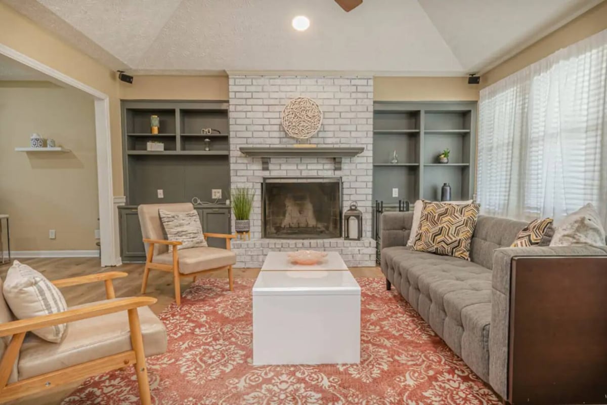 A cozy living room features a gray sofa paired with two wooden armchairs. A white coffee table is positioned on a decorative rug, while the stone fireplace is framed by built-in shelves. Natural light filters through sheer curtains, creating a warm, inviting space.