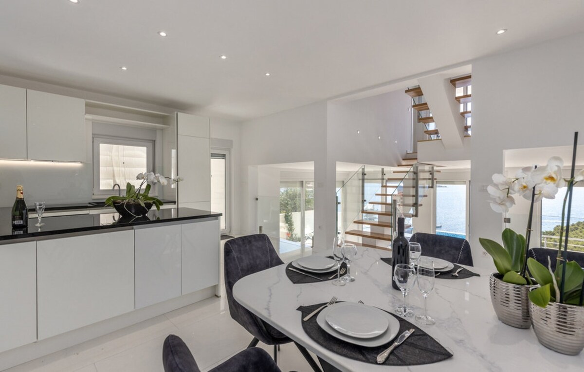 A modern kitchen and dining area is showcased, featuring a marble dining table set for six. The sleek cabinetry is complemented by stainless steel appliances. Large windows allow for natural light, illuminating the space and offering views of the sea and surrounding landscape.