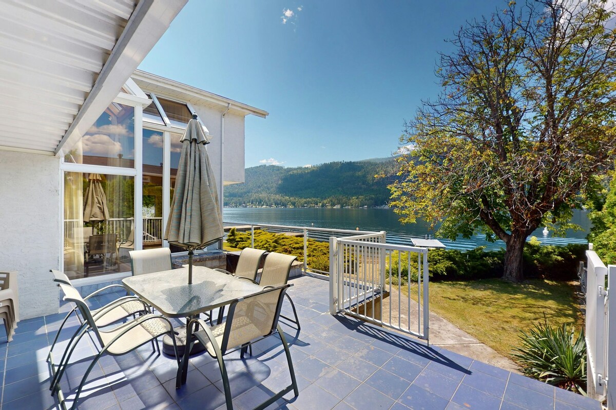 An outdoor patio area features a glass-top table surrounded by four metal chairs, set against the backdrop of serene lake views and lush greenery. A large umbrella provides shade, and a vibrant tree casts a gentle shadow over the space.