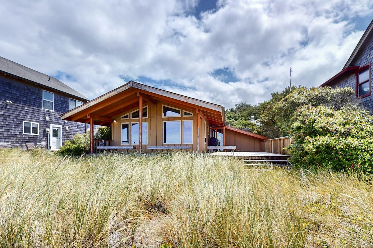A charming beach house is shown amid swaying coastal grass, with a covered deck providing an inviting outdoor space. Large windows reflect the sky, allowing natural light to fill the interior. Nearby structures are visible, set against a backdrop of gentle cloud formations.