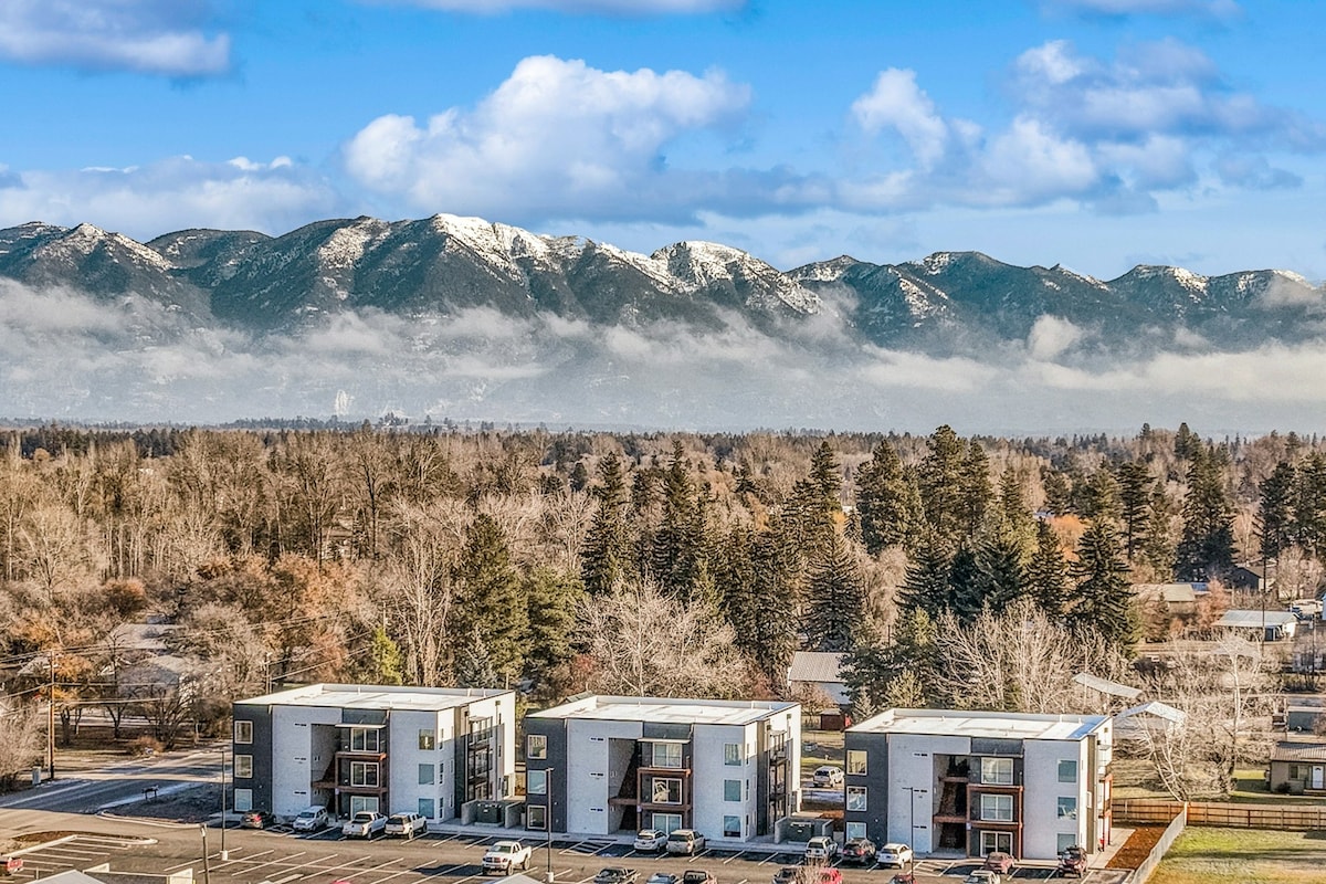An aerial view captures modern apartment buildings surrounded by lush trees, with a parking area in the foreground. The snow-capped mountains rise majestically in the background under a clear blue sky, enhancing the natural beauty of the setting.