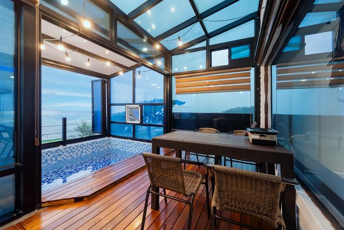 An enclosed terrace features a private swimming pool with azure tiles and wooden decking. A dining table with wicker chairs is present, complementing the serene ocean view visible through the large glass panels. Warm string lights provide soft illumination, enhancing the tranquil setting.