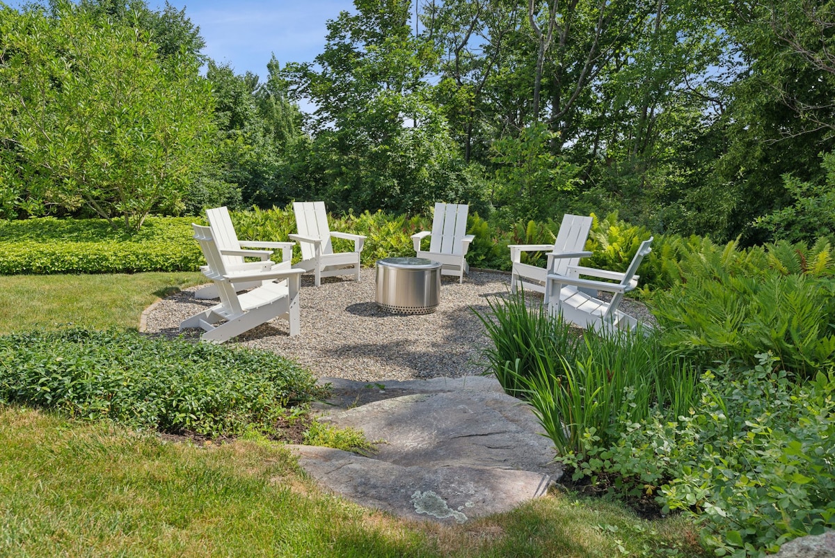 An outdoor firepit area is surrounded by four white Adirondack chairs arranged in a circular pattern. Lush greenery and perennial plants are visible in the background, while a smooth stone path leads to the gravel area, providing a serene setting for gatherings.