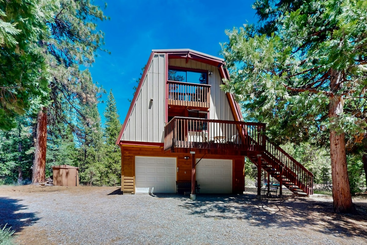 A multi-level cabin features a sloped roof and a raised deck, surrounded by tall pine trees. The entryway has a double garage below, with a gravel driveway leading to the front. Outdoor space is offered, inviting connection with nature.