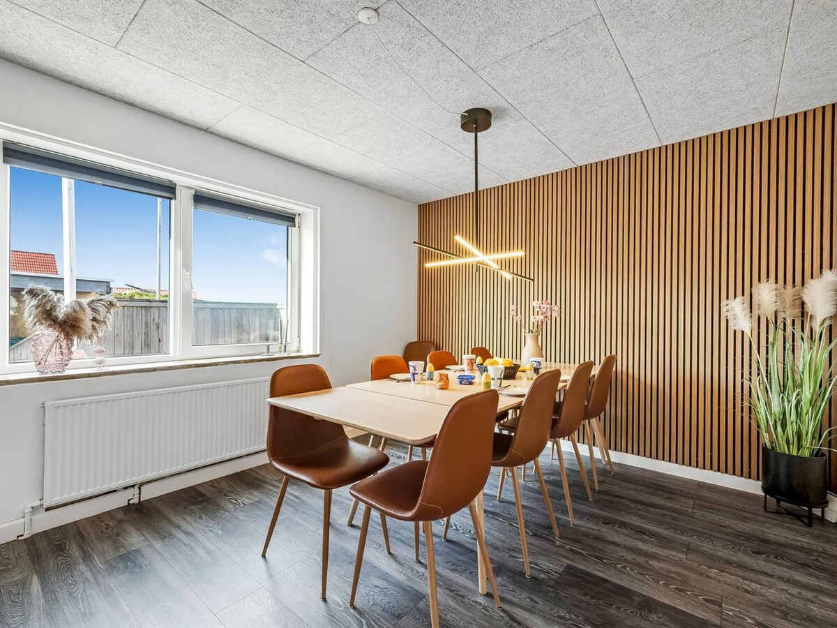 A dining area features a large table surrounded by eight chairs. Natural light filters through a large window, with wooden paneling on one wall enhancing the space. A plant in the corner adds a touch of greenery, creating a bright and welcoming atmosphere.