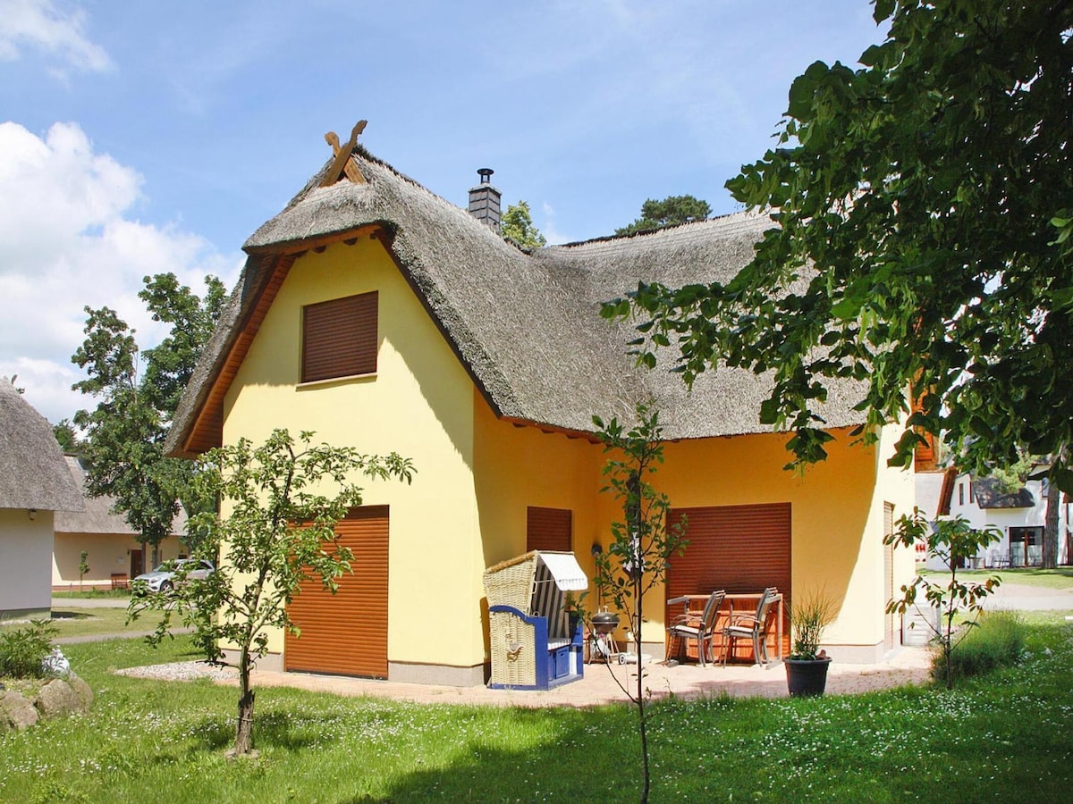A charming thatched cottage is depicted, featuring a yellow exterior and a traditional roofline. The building is surrounded by green foliage and a well-maintained garden area, with a seating arrangement visible on the outdoor patio. The overall setting provides a serene, natural ambiance.