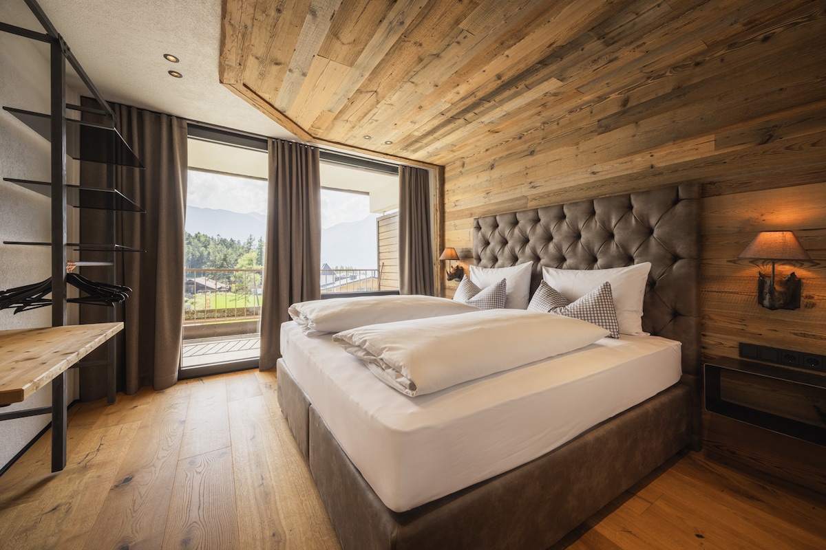 A spacious bedroom features a comfortable double bed dressed with crisp white linens, complemented by an elegant headboard. Natural light fills the room through large glass doors that open to a balcony, showcasing scenic views. The warm wood accents create a cozy atmosphere.