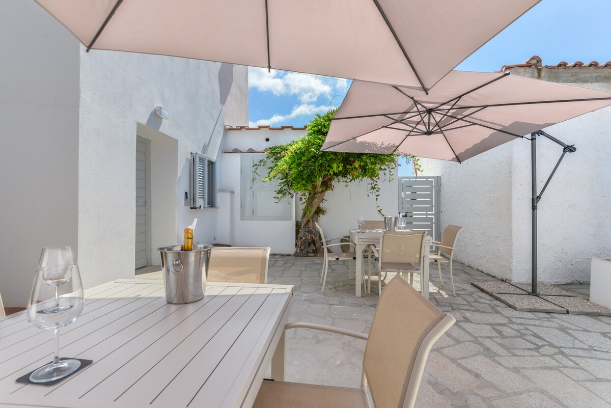 An outdoor patio area is shown, featuring a dining table with light-colored chairs. Two large umbrellas provide shade, alongside a small table with a wine cooler. A well-maintained tree adds greenery, with a versatile space designed for relaxation or dining.
