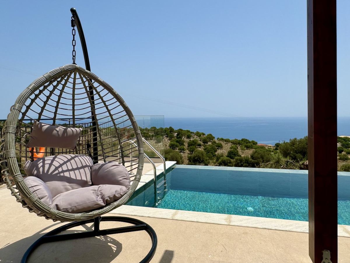 A circular hanging chair with soft cushions is positioned near the infinity pool, offering panoramic views of the blue sea. Lush greenery lines the horizon, while the pool's water reflects the clear sky. The scene suggests a serene outdoor space for relaxation.