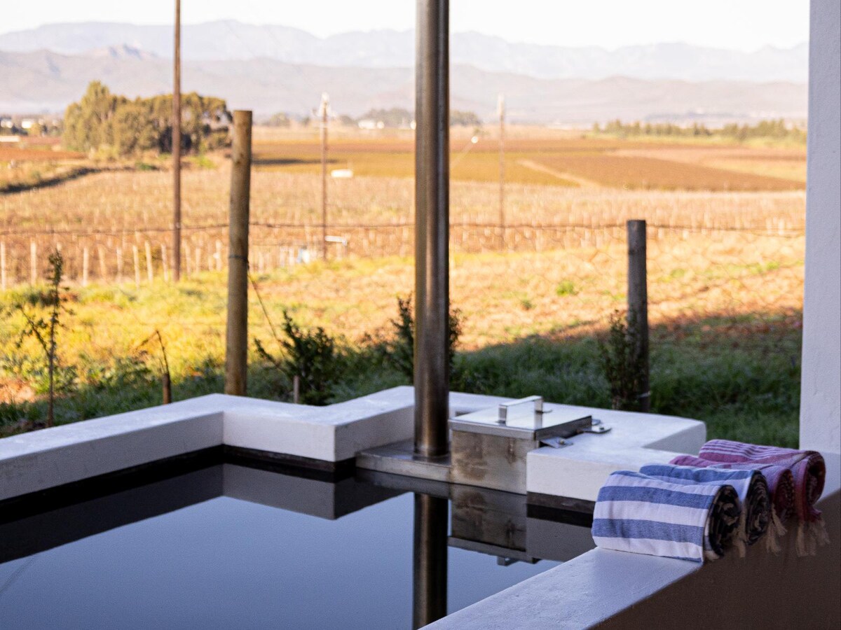 A serene wood-fired hot tub is positioned on a patio, surrounded by lush greenery, with a view of expansive vineyards and rolling hills in the background. Neatly arranged towels in various colors rest on the edge, while a chimney provides warmth.