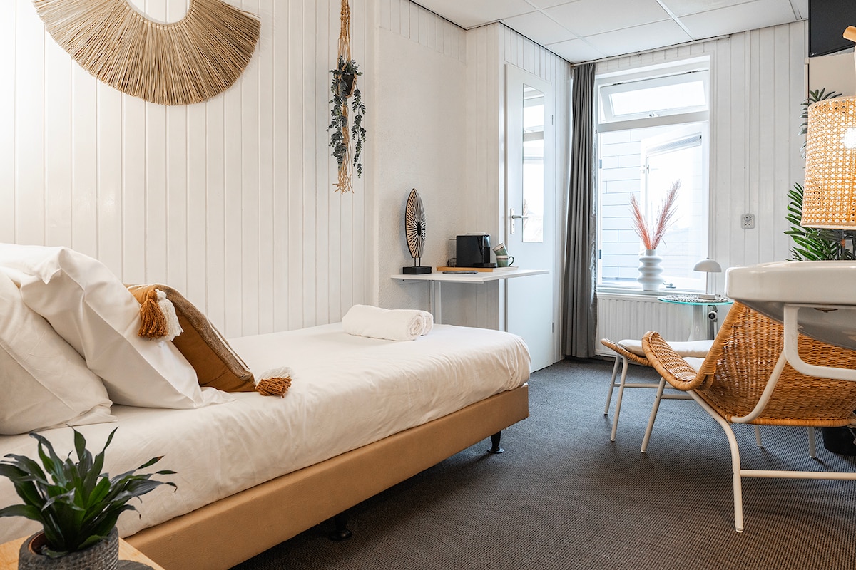 A compact room features a comfortable boxspring bed dressed in crisp linens. A small desk and chair are positioned for convenience, while a cozy lounge chair is situated nearby. Light filters in through a large window, offering a serene atmosphere complemented by modern decor and greenery.
