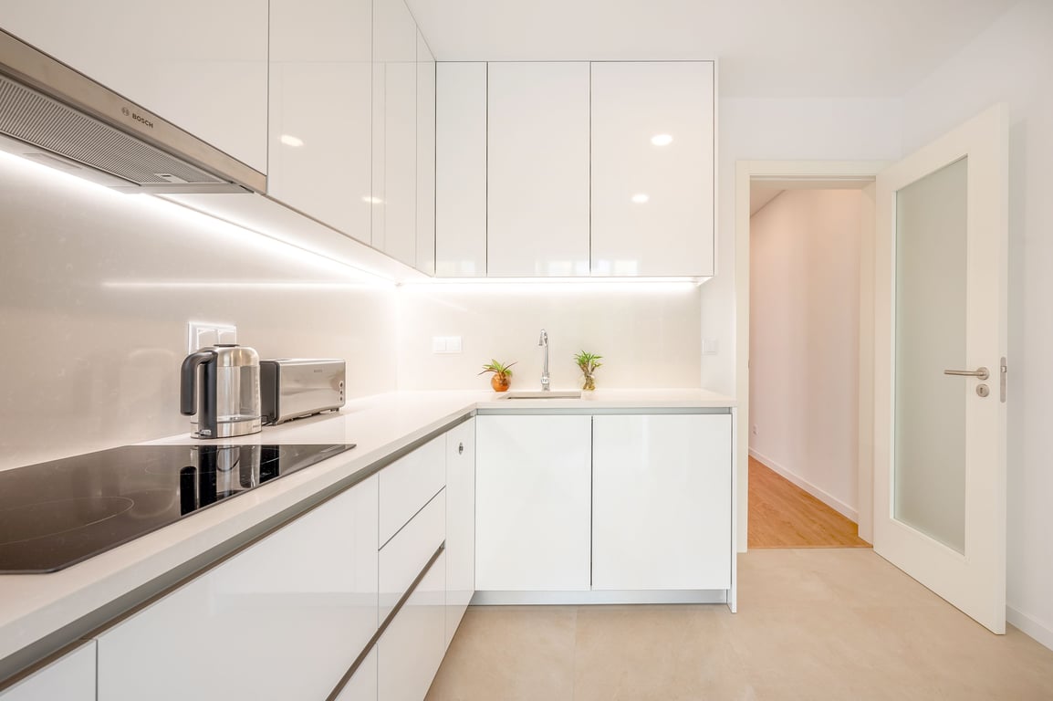A modern kitchen is shown with sleek white cabinetry and a clean countertop. The space is equipped with essential appliances, including a cooking plate and microwave. Even lighting enhances the bright atmosphere, while a small plant adds a touch of greenery near the sink.