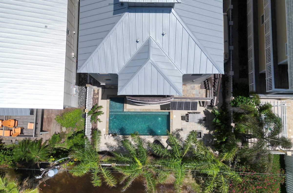 An aerial view of a three-level home surrounded by tropical landscaping. A private heated pool is centered in the backyard, with lounge chairs and shaded areas. The home's modern architecture is showcased with a metal roof and a spacious outdoor area accessible from multiple levels.